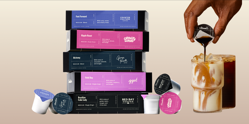 Stack of coffee boxes with pods beside a hand pouring coffee creamer into an iced latte glass