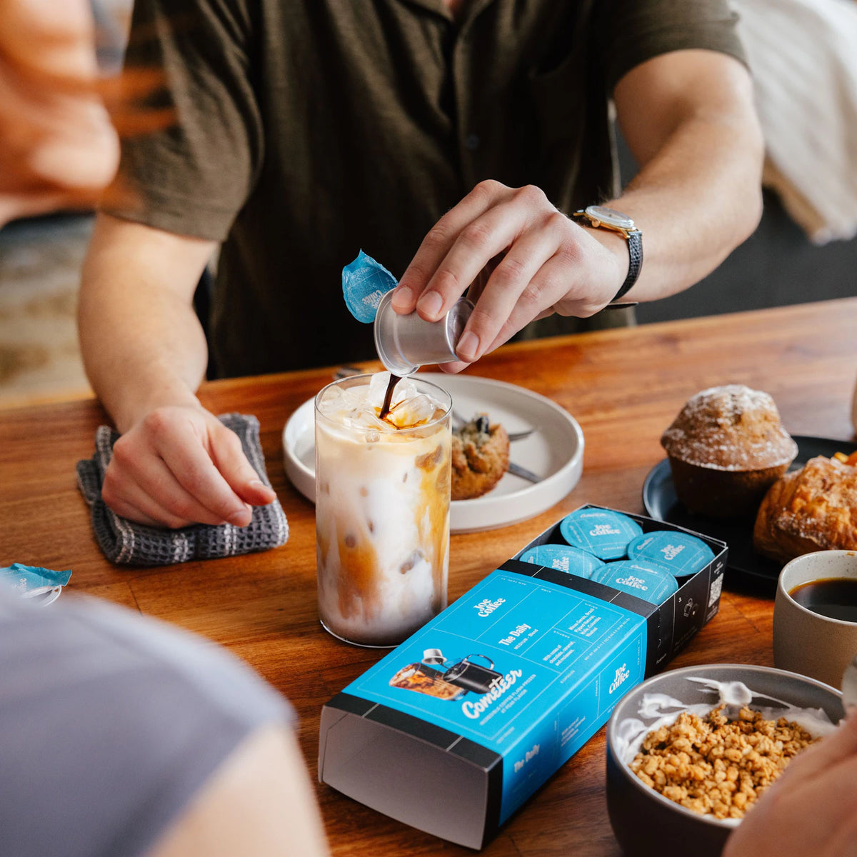 A Cometeer capsule being poured into an iced latte beside a blue coffee sleeve and breakfast.