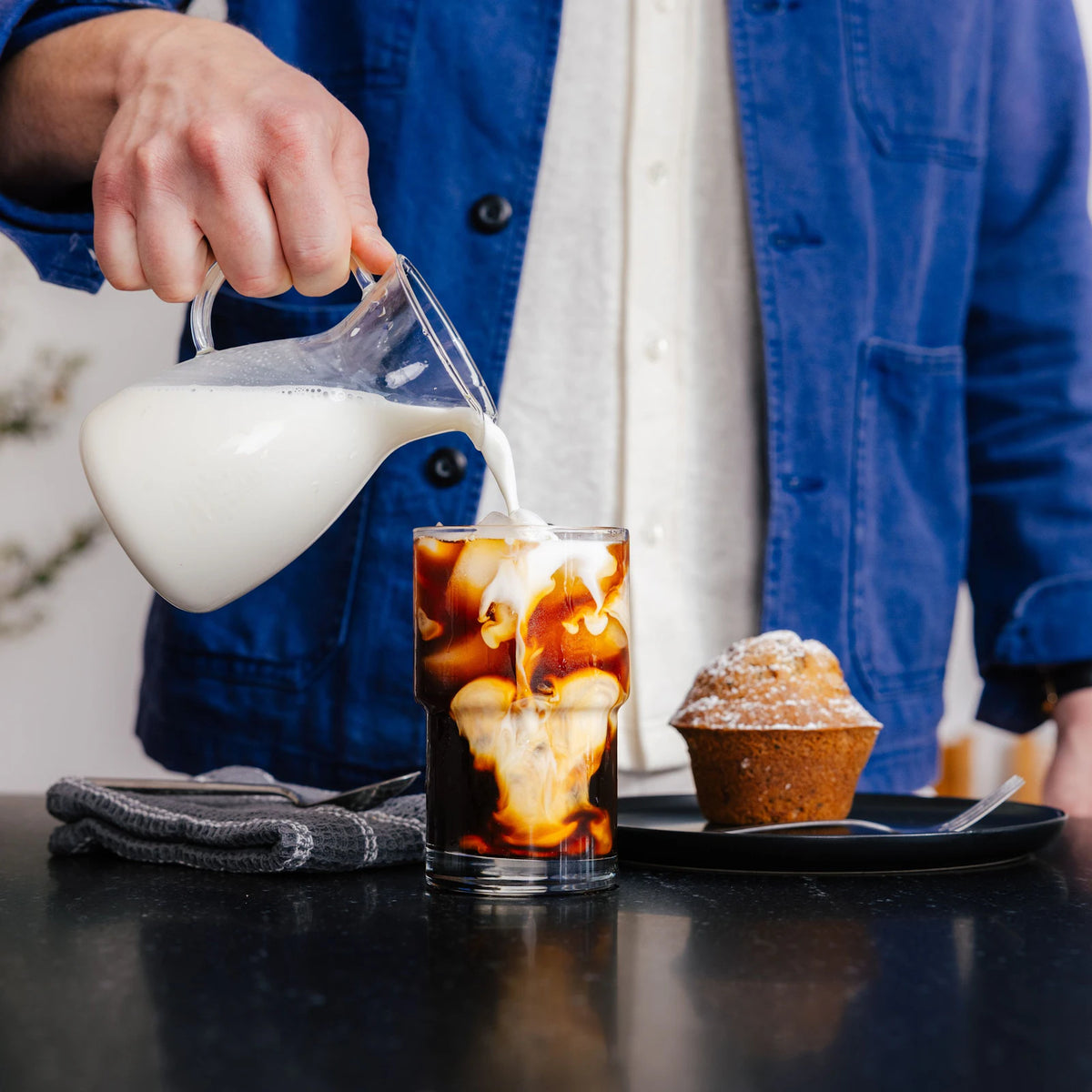 Milk being poured into a glass of iced coffee beside a muffin.